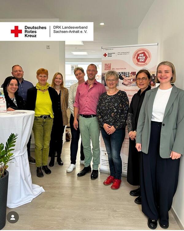 Acht Personen stehen in einem Halbkreis vor einen Roll-up mit einem runden Logo des Enagementzentrums Sangerhausen, oben links ist das DRK Logo des DRK Landesverbandes vor weißem Hintergrund zu sehen.