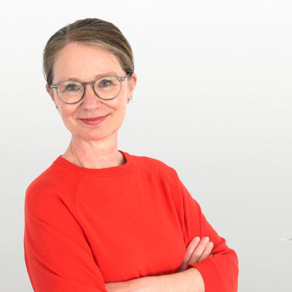 Eine Frau mit Brille steht mit Blick in die Kamera lächeln da. Sie trägt einen rotem Pullover und hat die Arme verschrenkt.