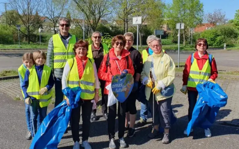 eine Gruppe an älteren Menschen und 2 kleinen Kindern, die Warnwesten tragen, sowie Mülltüten in der Hand halten