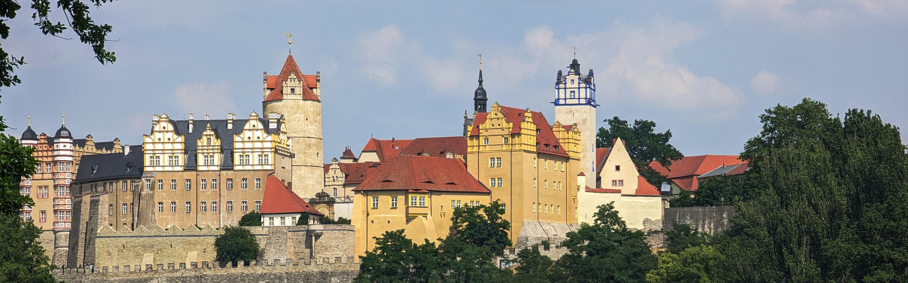 Auf dem Bild ist eine Burg abgebildet. Es sind Bäume auf der rechten Seite des Bildes zu sehen die am Ufer wachsen. 
