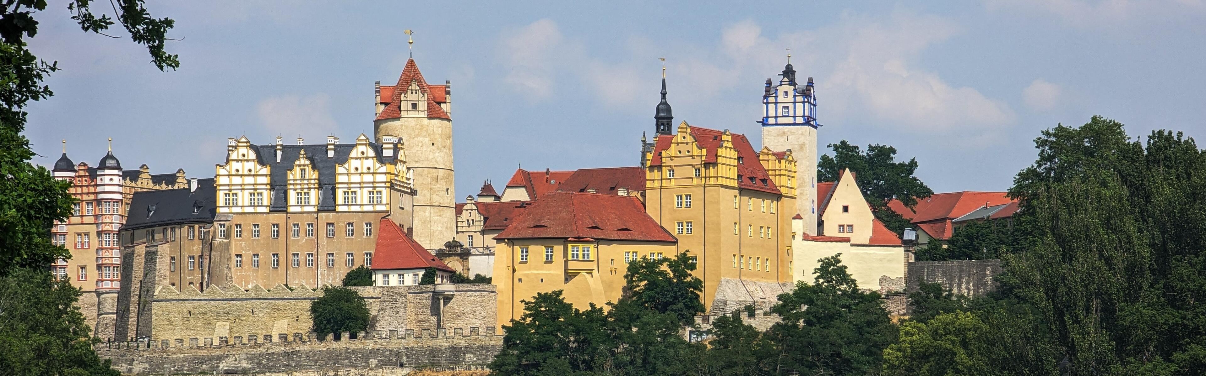 Auf dem Bild ist eine Burg abgebildet. Es sind Bäume auf der rechten Seite des Bildes zu sehen die am Ufer wachsen. 
