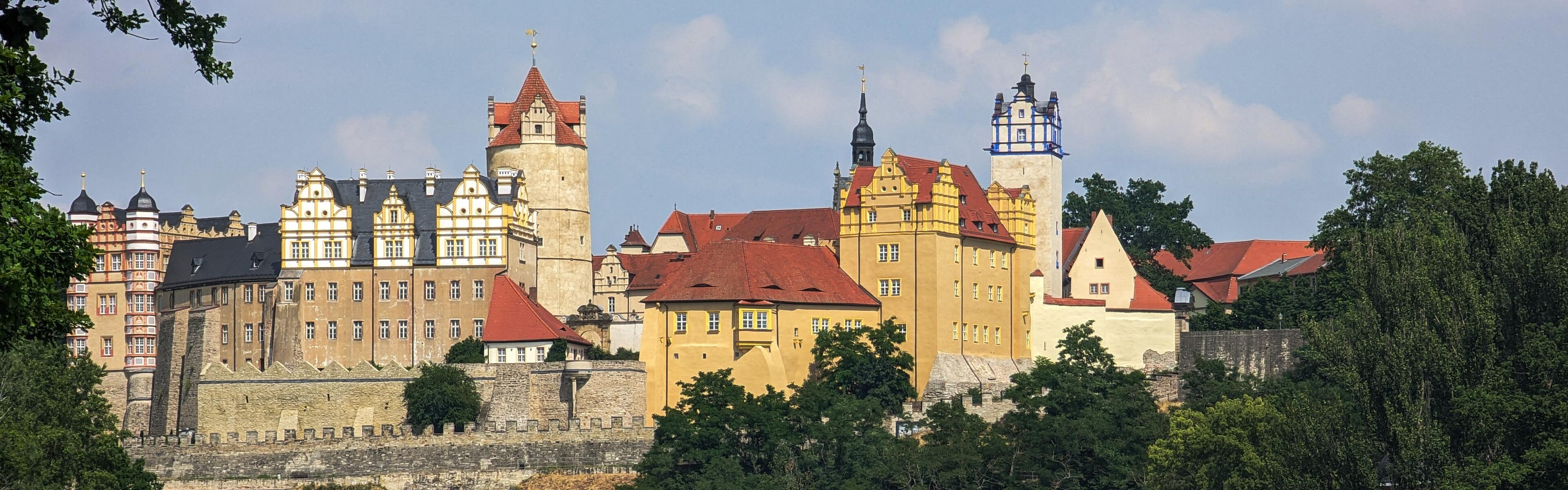 Auf dem Bild ist eine Burg abgebildet. Es sind Bäume auf der rechten Seite des Bildes zu sehen die am Ufer wachsen. 