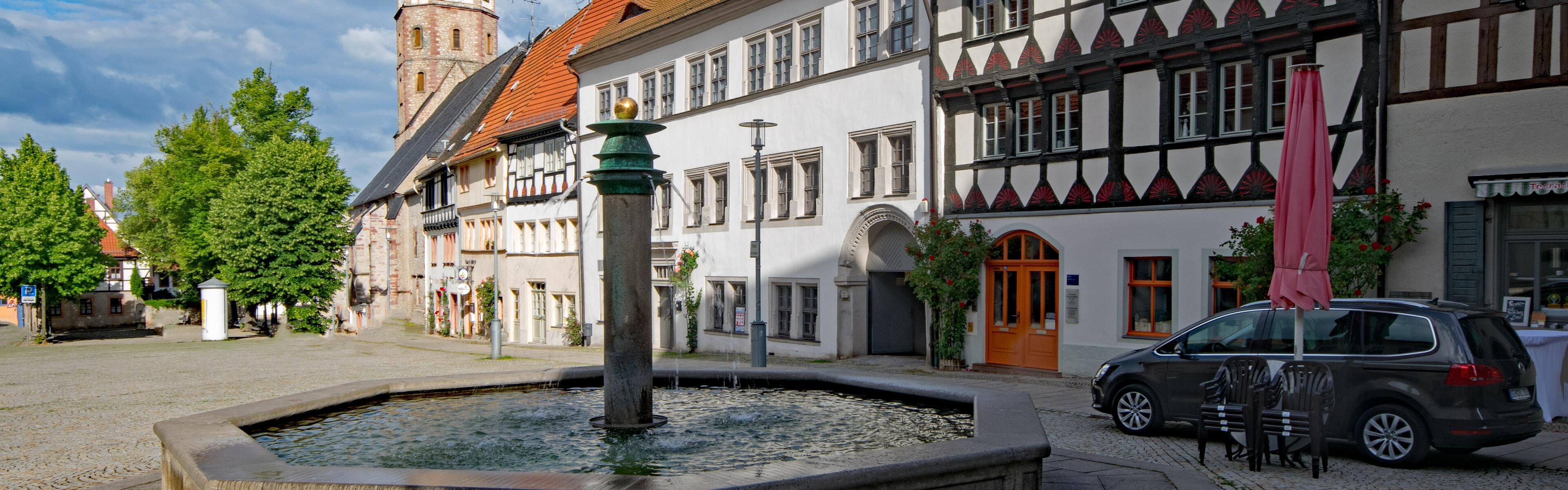 Im Vordergrund steht ein Brunnen, dahinter befinden sich Fachwerkhäuser. Im Hintergrund, mittig im Bild, sieht man den Turm einer Kirche. 