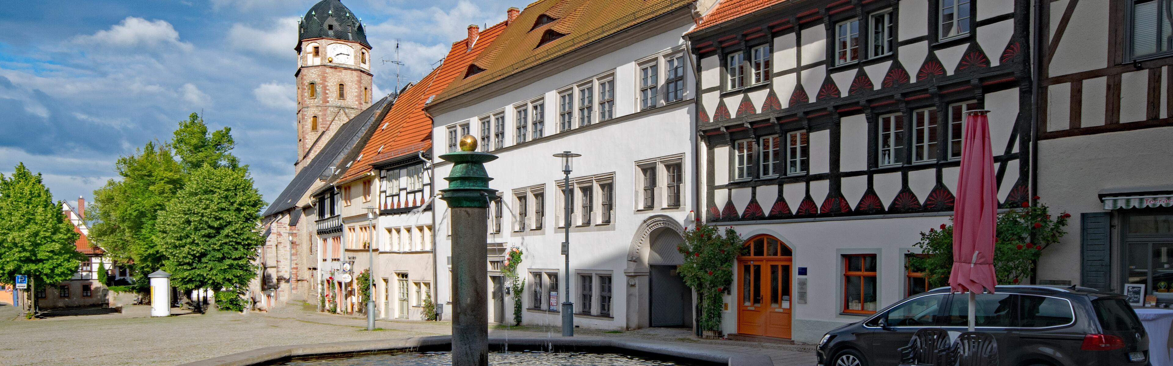 Im Vordergrund steht ein Brunnen, dahinter befinden sich Fachwerkhäuser. Im Hintergrund, mittig im Bild, sieht man den Turm einer Kirche. 