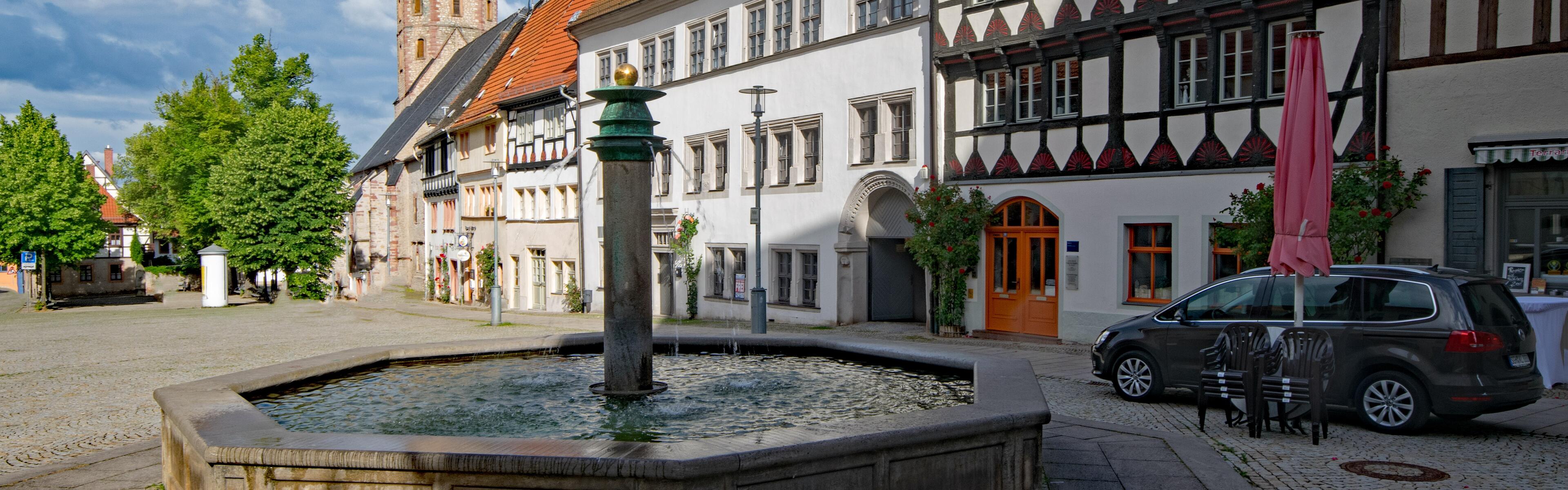 Im Vordergrund steht ein Brunnen, dahinter befinden sich Fachwerkhäuser. Im Hintergrund, mittig im Bild, sieht man den Turm einer Kirche. 