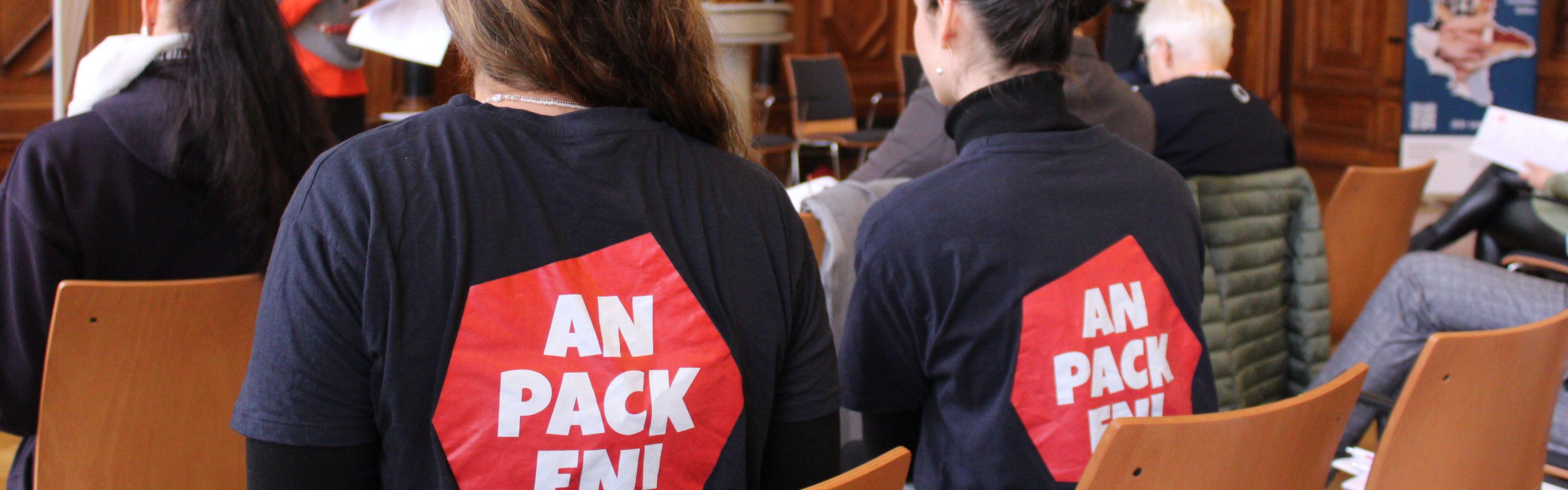 Zwei Frauen sitzen mit dem Rücken zur Kamera in einem festlichen Saal, sie tragen beide blaue T-Shirts auf denen ein rotes Sechsecklogo zu sehen ist mit der Inschrift Anpacken!