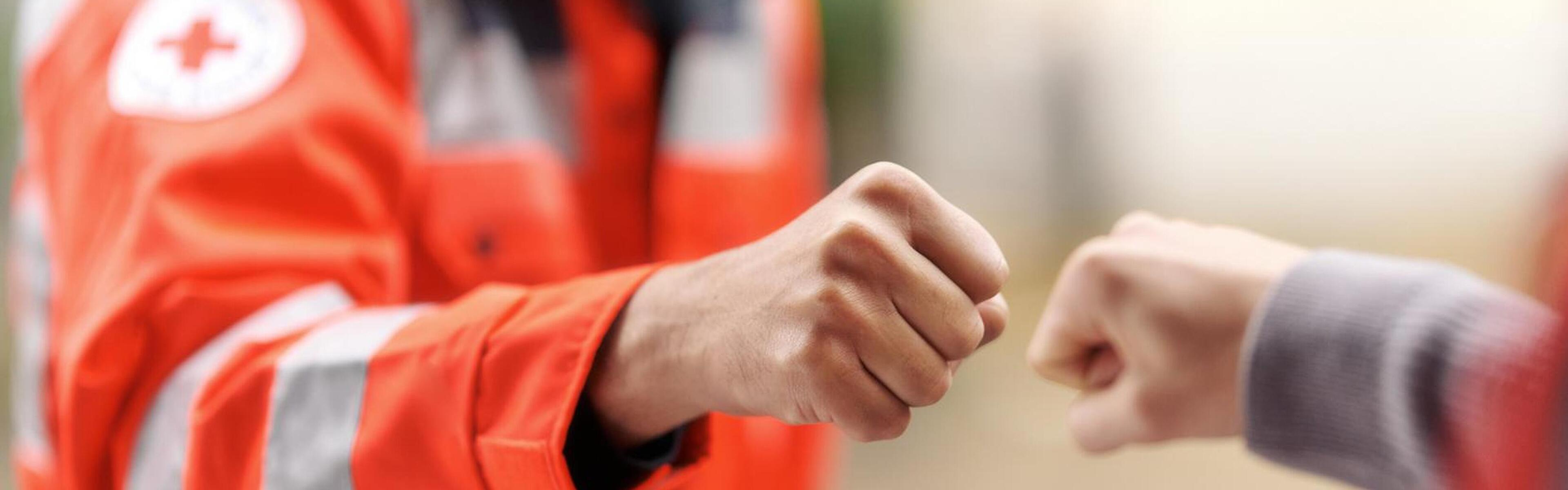 Zwei Menschen geben sich ein Faustgruß, bei der linken Person ist sichtbar, dass sie eine DRK Einsatzjacke in Neonorange trägt.